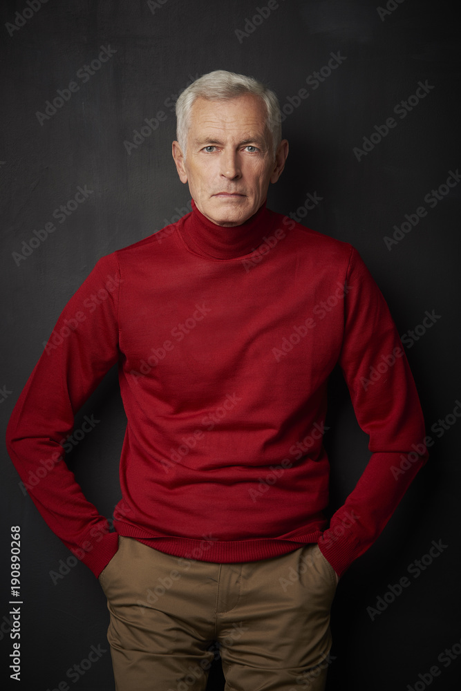 Casual senior man portrait. Handsome elderly man wearing red turtle neck sweater and looking at camera while standing against at dark background.
