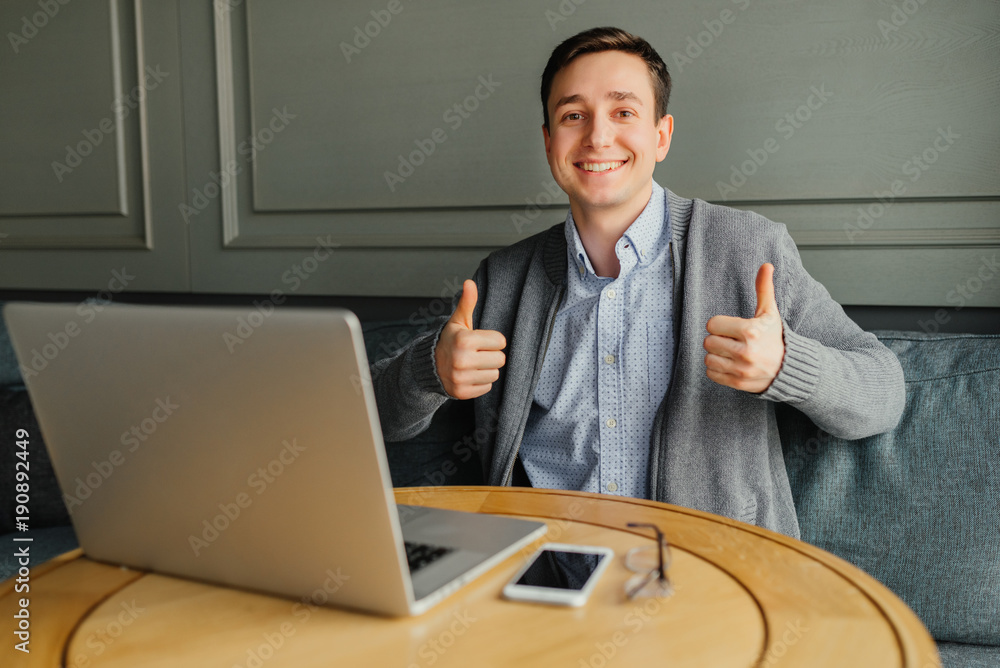 Happy smiling successful young man sitting with laptop in cafe and shows gesture thumbs up