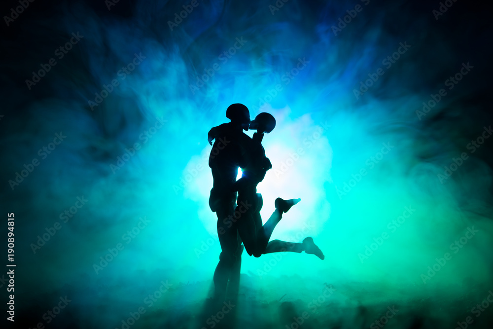 Love Valentine`s Day concept.Sillhouette of sweet young couple in love standing in the field and hugging on dark toned foggy background. Decoration with doll figures on table shot.