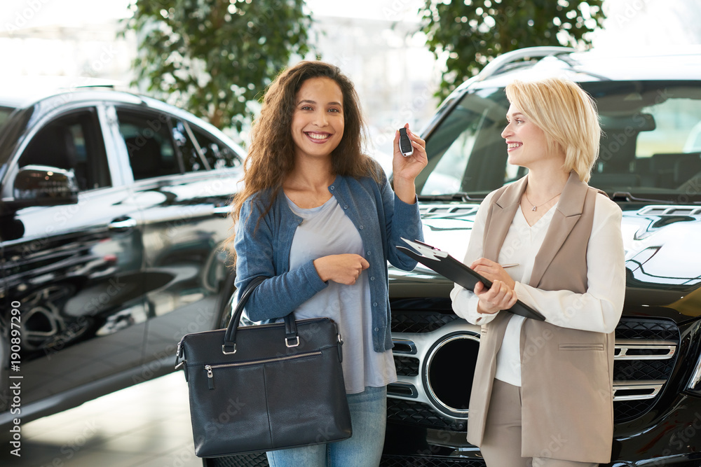 Attractive young customer showing key of new car to camera and posing for photography with toothy smile while blonde saleswoman standing next to her, interior of modern showroom on background Attractive young customer showing key of new car to camera and posing for photography with toothy smile while blonde saleswoman standing next to her, interior of modern showroom on background