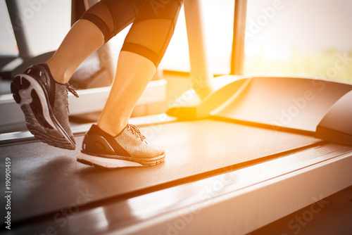Lower body at legs part of Fitness girl running on running machine or treadmill in fitness gym with sun ray. Warm tone. Healthy and Exercise activity concept. Workout and Strength training theme.
