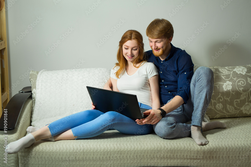 beautiful young couple guy with a girl with red hair sitting on the couch with a laptop