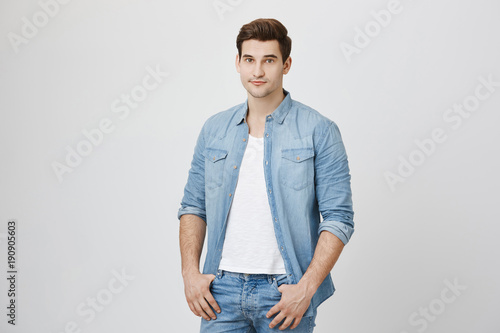 Portrait of handsome young man with confident look, holding thumbs in pockets while looking at camera, isolated over white wall. New teacher introduces in front of students © Cookie Studio