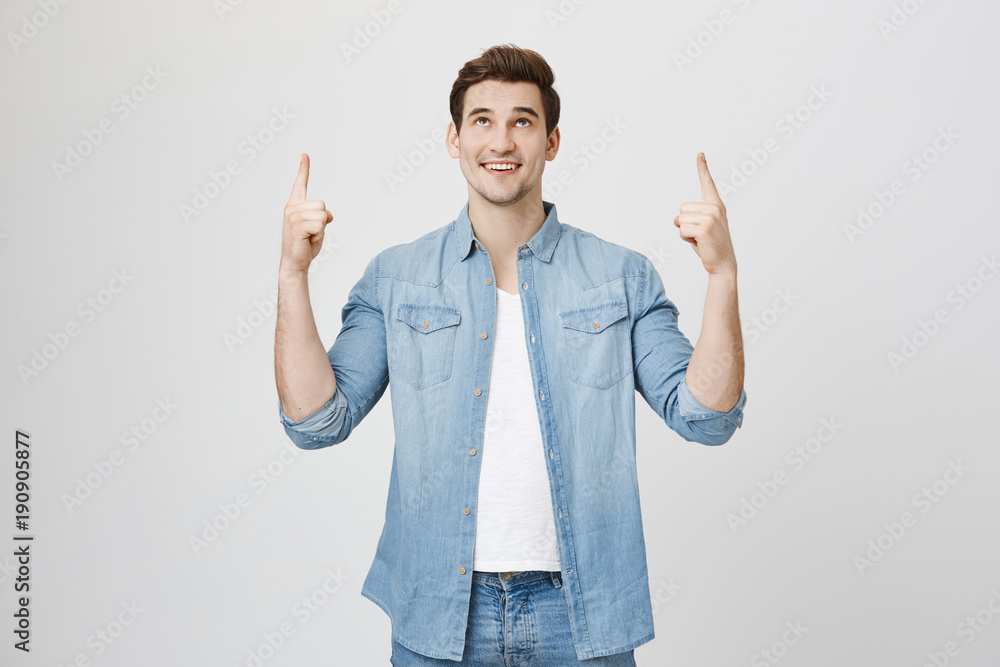 Happy pleased Caucasian male with stubble points with forefingers upwards dressed in denim clothes, smiles joyfully, indicates at gray wall with copy space for advertisement or promotional text. Happy pleased Caucasian male with stubble points with forefingers upwards dressed in denim clothes, smiles joyfully, indicates at gray wall with copy space for advertisement or promotional text.