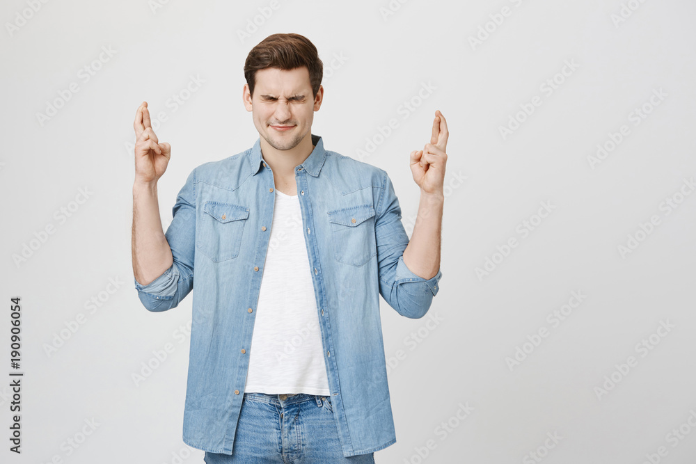 Studio portrait of young freelancer in denim clothes standing with closed eyes and showing praying gesture with crossed fingers, over gray background. Student begs for canceling exam Studio portrait of young freelancer in denim clothes standing with closed eyes and showing praying gesture with crossed fingers, over gray background. Student begs for canceling exam