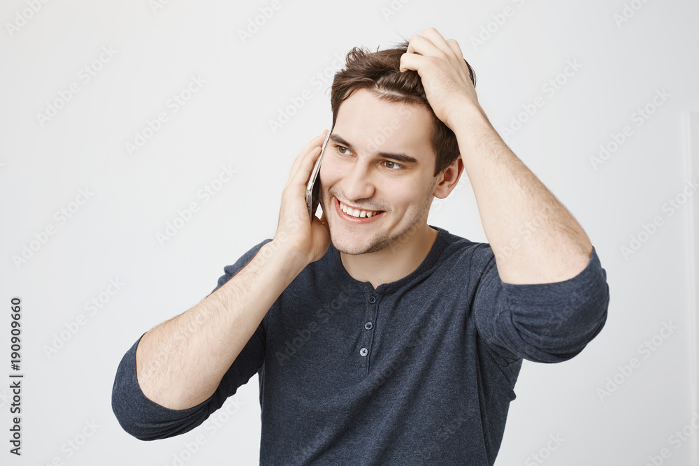 Indoor portrait of handsome young sportsman talking on smartphone while touching his hair and looking aside, standing over gray background. So, I was wondering if you want to go out sometimes Indoor portrait of handsome young sportsman talking on smartphone while touching his hair and looking aside, standing over gray background. So, I was wondering if you want to go out sometimes