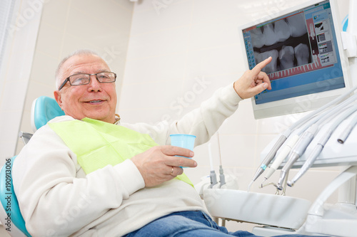 Old senior man in dental clinic. Sitting in a chair and look X-Ray on computer