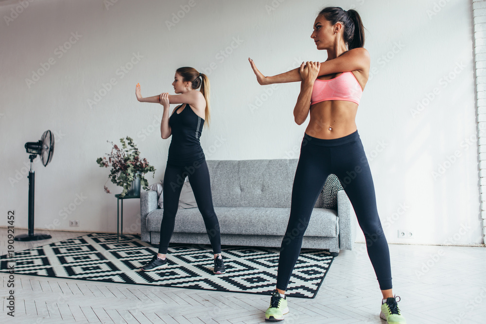Obraz premium Two fit young woman stretching their arms exercising at home. Caucasian female fitness models working out in the morning.