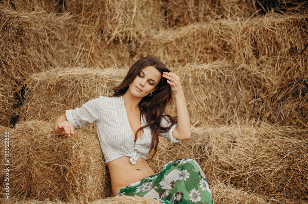 Thinking about love. Portrait of a long hair woman on a stack of straw. The concept of photo harvesting, fertility, beer. Woman in a white blouse and green skirt with field flowers.