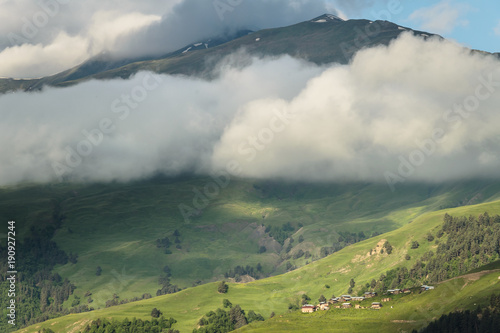 Early morning at Caucasus mountains, Georgia, Tusheti region. Small village located on the green hill on the foreground and high peak surrounded with clouds on the background.