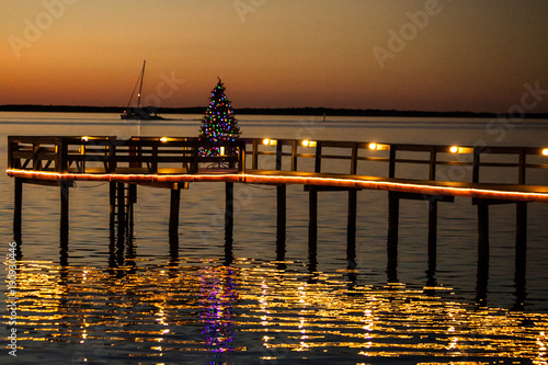 A Dunedin, Florida pier lit with Chrismas lights and a Christmas tree at sunset with a sailboat cruising by in the background.