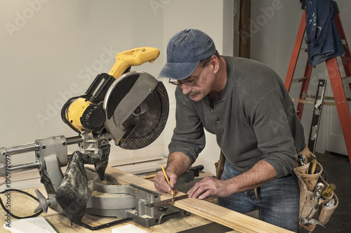 male carpenter wearing old tool belt, marking wood board with old square and pencil, next to electric chop saw, at fixer upper home residential construction site