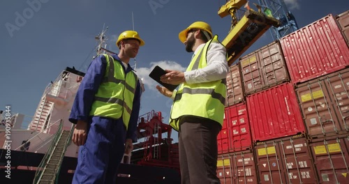 Dock workers discuss logistics at the harbor on a digital tablet amidst shipping industry activity. Shot on RED Epic.