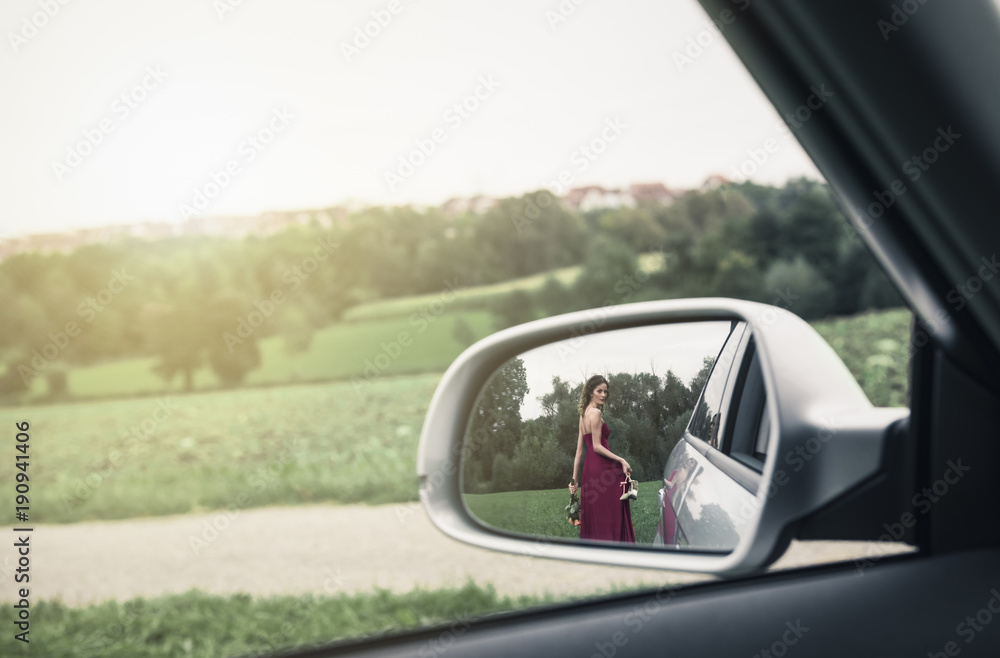 Elegant woman seen in the rear view mirror of car - Young woman, in an evening red dress, with her shoes and a flower bouquet in her hands, seen in the rear view mirror, walking away from the car.