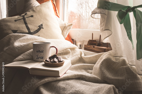 View of a book, coffee, chocolate, pillows and blanket by a window with a view of autumn or winter landscape