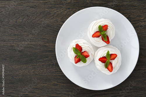 Meringue Pavlova desserts with strawberry slices and mint leaves in white plate on dark wooden background, top view