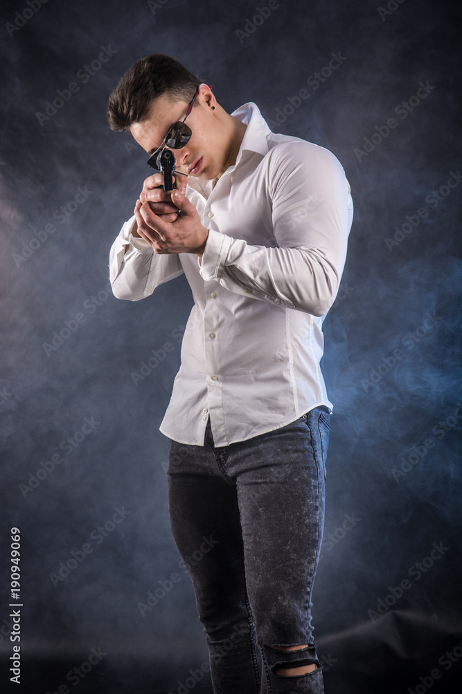 Young handsome man pointing gun, wearing black t-shirt, on dark background in studio Young handsome man pointing gun, wearing black t-shirt, on dark background in studio