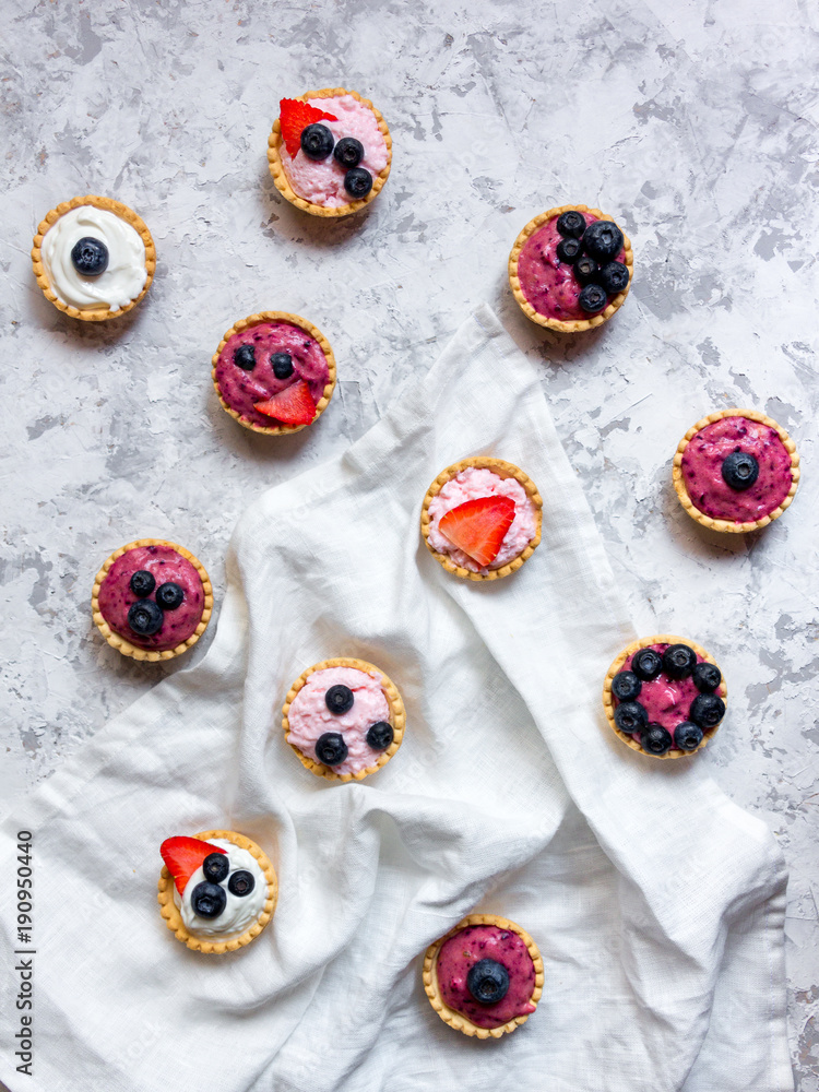 Overhead still life of sweet mini tartlets with cream and berries on white textured background. Simple dessert snack recipe with blueverry and strawberry. Summer food concept.