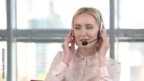 Cheerful lady working in call center. Business woman is talking with someone using headset, huge bright windows background.