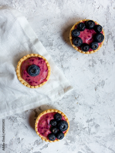 Overhead still life of sweet mini tartlets with cream and berries on white textured background. Simple dessert snack recipe with blueberry. Summer food concept.