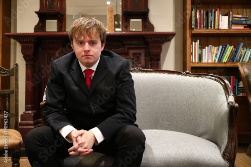 A young man looking serious in a suit sat on a sofa