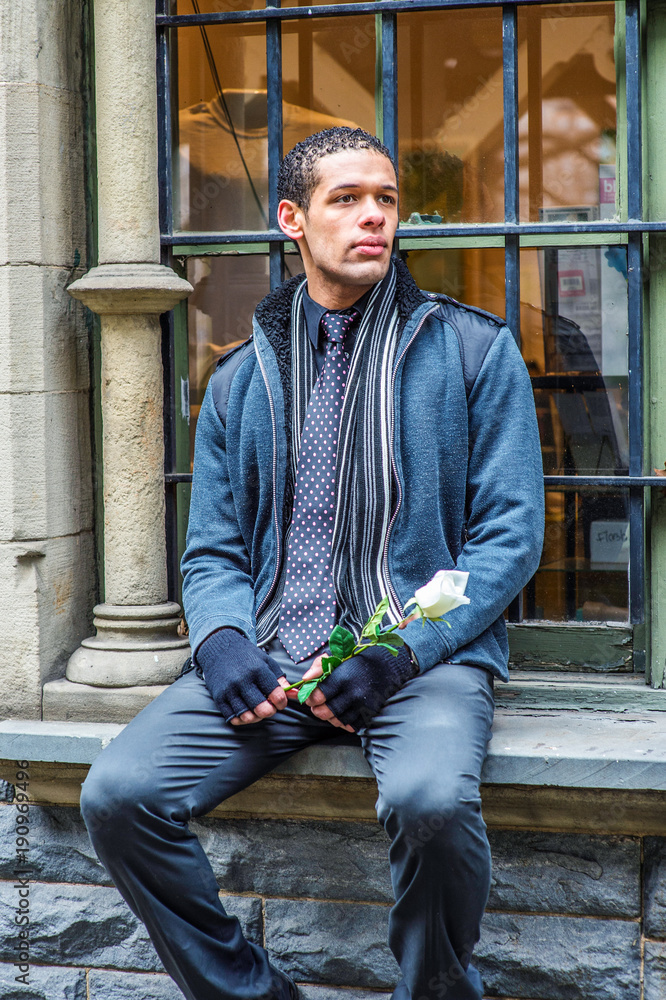 A young guy, holding a white rose and sitting on a window frame, is waiting for you..