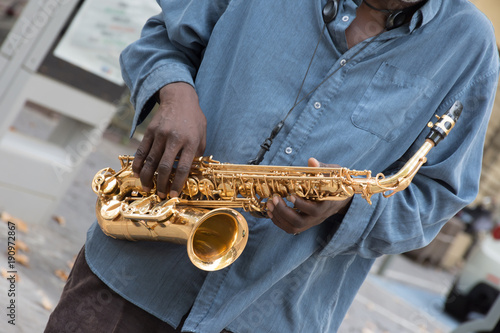 A black saxophone player wearing a denim shirt. He is holding the gold colored sax at an angle at chest level. Only his torso is seen.