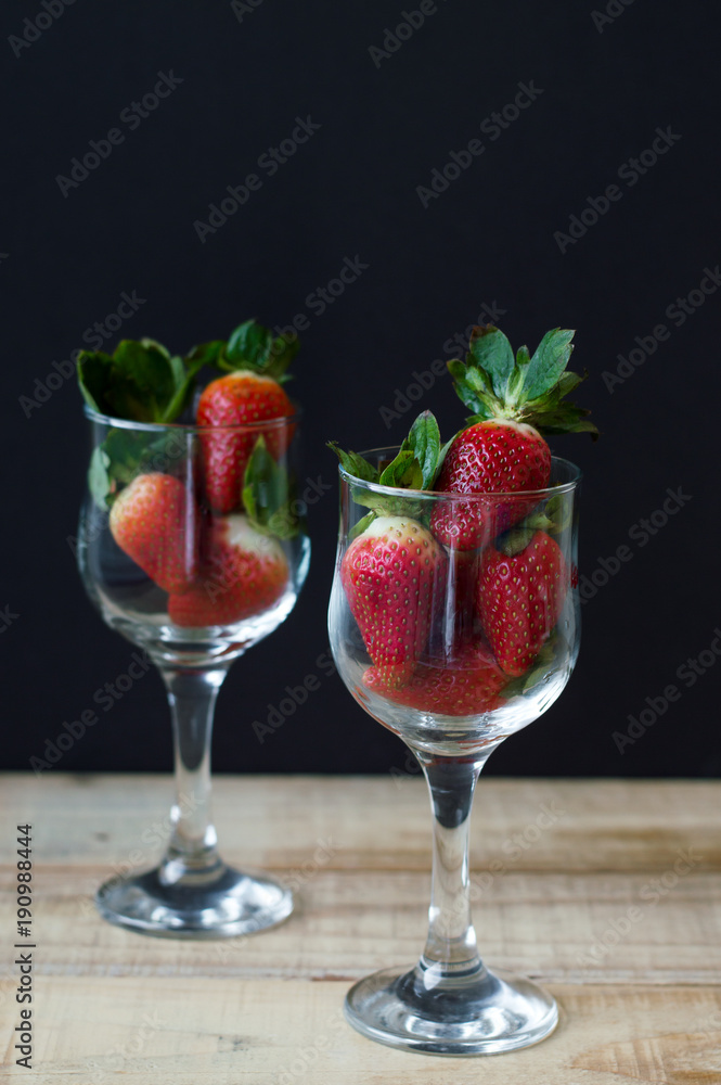Two vine glasses with fresh strawberry with green leaves on wooden table, black background. Healthy dessert Two vine glasses with fresh strawberry with green leaves on wooden table, black background. Healthy dessert