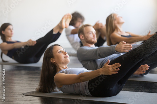Group of young sporty people practicing fitness lesson with instructor, doing warming up exercise, boat pose, working out, students training in club, friends at fitness lesson, indoor, studio