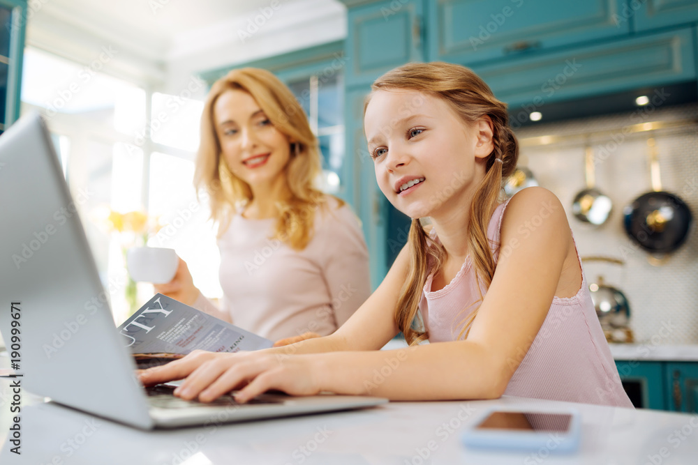 Happy at home. Beautiful inspired blond girl smiling and working on the laptop while her mother drinking coffee Happy at home. Beautiful inspired blond girl smiling and working on the laptop while her mother drinking coffee