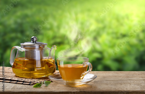 Warm cup and glass jugs or jars of tea and tea leaf and sack on the wooden table and the tea plantations background