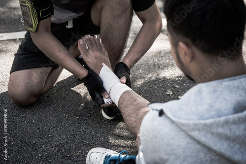 A man uses elastic bandage for pain relief one man's ankle after a workout in a park.