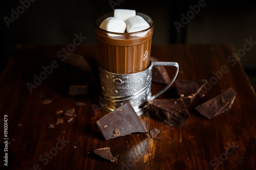 An underexposed horizontal image of hot chocolate in a glass in a metal glass-holder, decorated with marshmallows and pieces of dark chocolate on a wooden table. Selective focus.