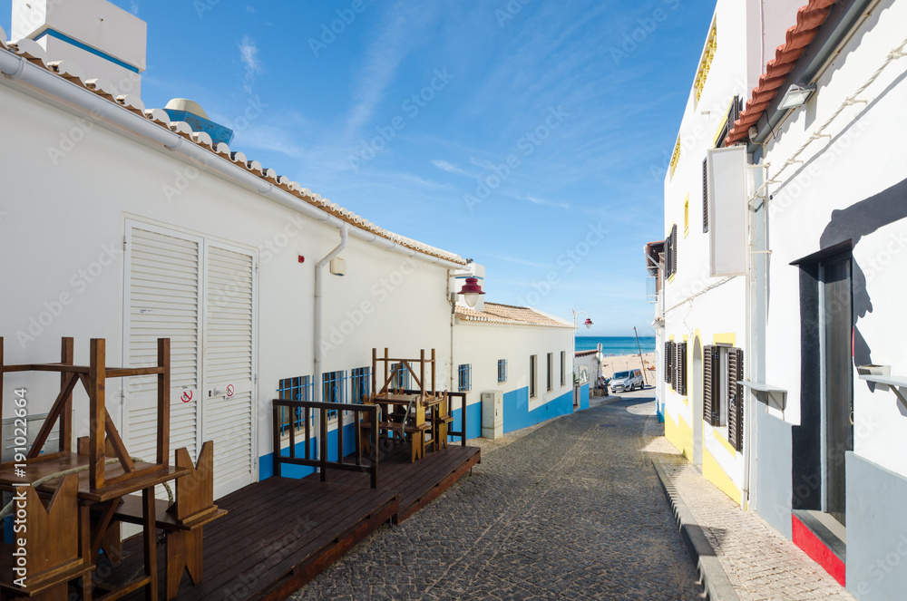 Narrow street of Salema village. Ocean view and whitewashed houses of the small authentic fishing village with a lots of small cafes, bars and sea food restaurants. Faro, Algarve, Southern Portugal.
