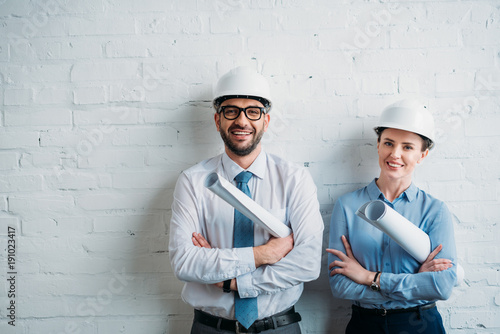 happy architects in hard hats standing in front of white brick wall with blueprints