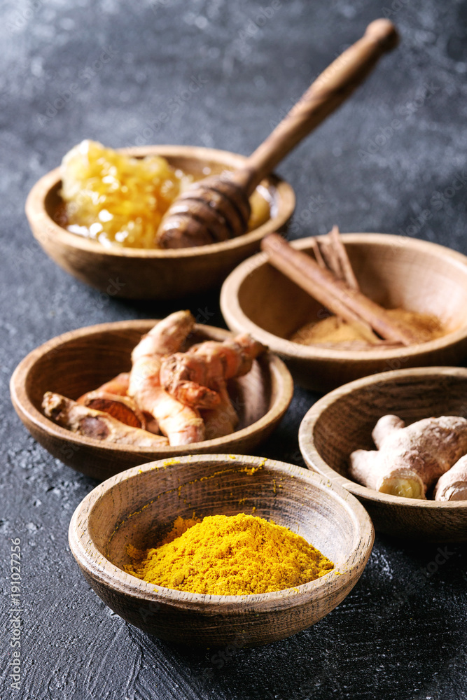 Fototapeta premium Ingredients for turmeric latte. Ground turmeric, curcuma root, cinnamon, ginger, honeycombs in wooden bowls over black texture background. Close up