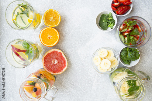 Glasses of detox water with sliced fruit, vegetables, berries and herbs and bowls with ingredients: strawberry, mint, spinach, grapefruit on a light stone background, top view