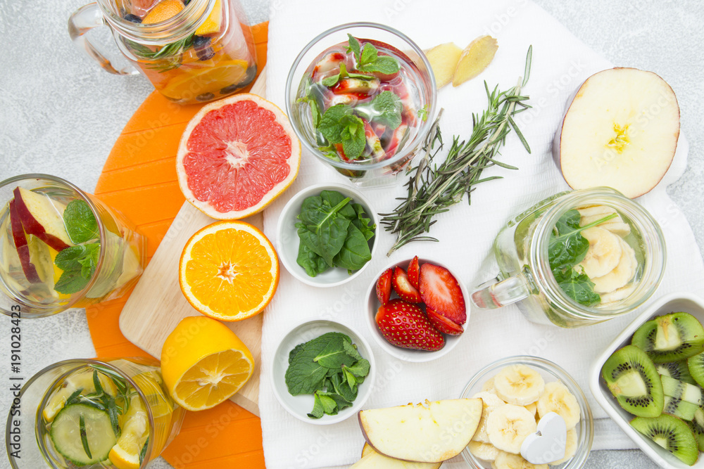 Glasses of detox water with sliced fruit, vegetables, berries and herbs, set of ingredients: banana, strawberry, orange, grapefruit, mint, spinach and rosemary on cutting boards and napkin, top view Glasses of detox water with sliced fruit, vegetables, berries and herbs, set of ingredients: banana, strawberry, orange, grapefruit, mint, spinach and rosemary on cutting boards and napkin, top view