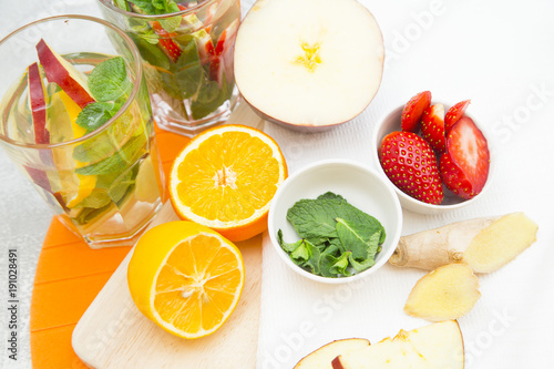Detox infused water with slices of fruit and herbs.Bowls with strawberry and spinach, orange and lemon cut in halves, sliced apple and ginger on cutting board and white napkin, top view