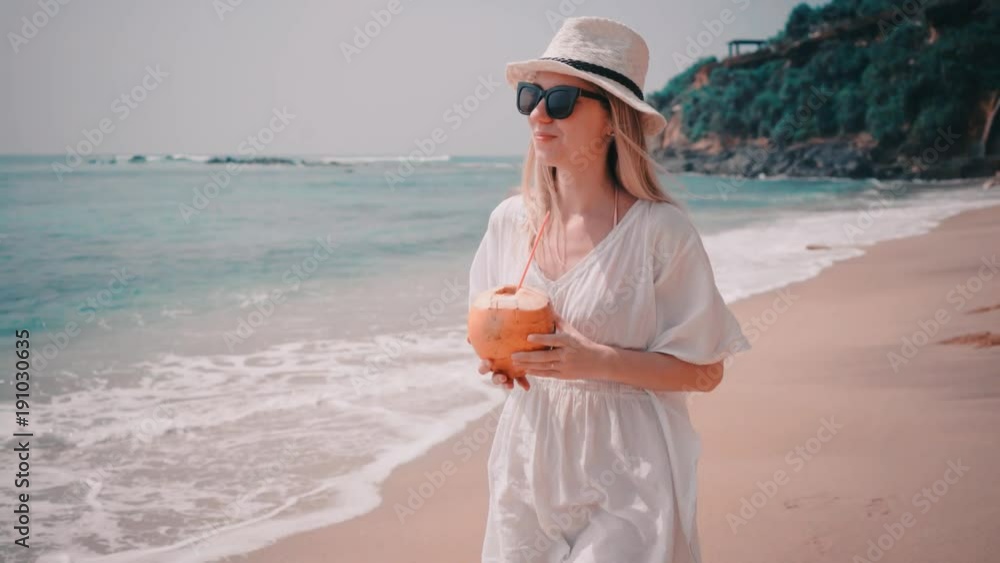 Woman drinks coconut milk and wallking on the ocean beach under blue sky in sunny day. Seascape view with palms. Woman drinks coconut milk and wallking on the ocean beach under blue sky in sunny day. Seascape view with palms.