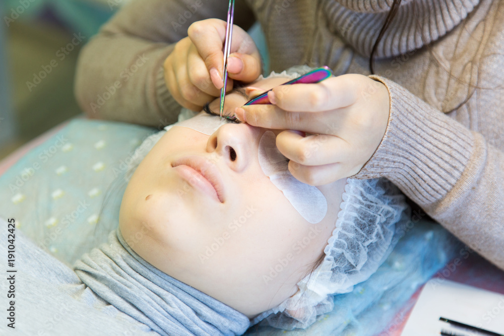 Volumetric eyelash extends in the beauty salon. The procedure for increasing eyelashes in front of a blonde girl is done by a master in a beauty salon