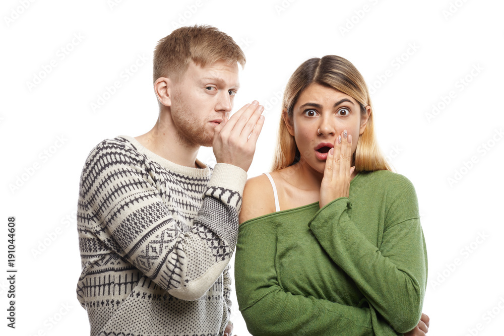 Attractive young unshaven male sharing secret or whispering gossips into his astonished girlfriend's ear, who is staring at camera with mouth wide opened, shocked with unexpected information Attractive young unshaven male sharing secret or whispering gossips into his astonished girlfriend's ear, who is staring at camera with mouth wide opened, shocked with unexpected information