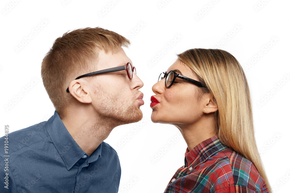 Profile studio portrait of beautiful young blonde lady wearing stylish eyeglasses and red lipstick standing in front of bearded geeky guy, both pouting lips and closing eyes for kissing each other Profile studio portrait of beautiful young blonde lady wearing stylish eyeglasses and red lipstick standing in front of bearded geeky guy, both pouting lips and closing eyes for kissing each other