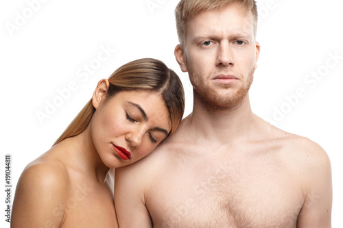 Sensual passionate adult couple posing topless in studio: handsome unshaven male looking at camera with serious expression while blonde woman keeping eyes closed and resting head on his shoulder