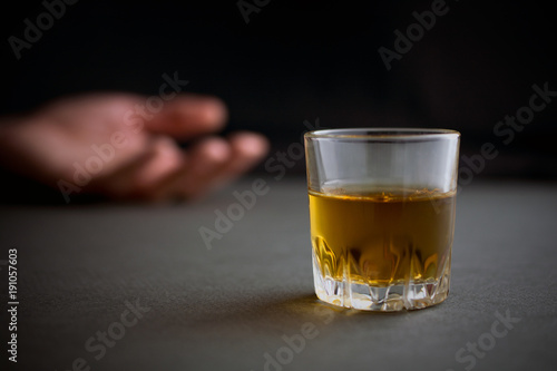 glass of whiskey or cognac or alcohol drink, hand of a drunk man in the background, alcoholism and alcohol abuse concept, defocused, selective focus, close up, gray table, dark background