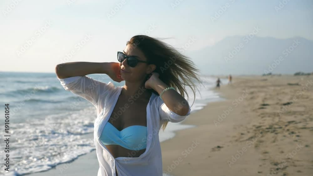 Portrait of beautiful girl in bikini and shirt walking along seashore and playing with her blonde hair. Happy young woman in sunglasses smiling and enjoying life during vacation. Travel concept