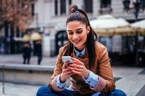 Smiling young woman using smartphone sitting at city street.