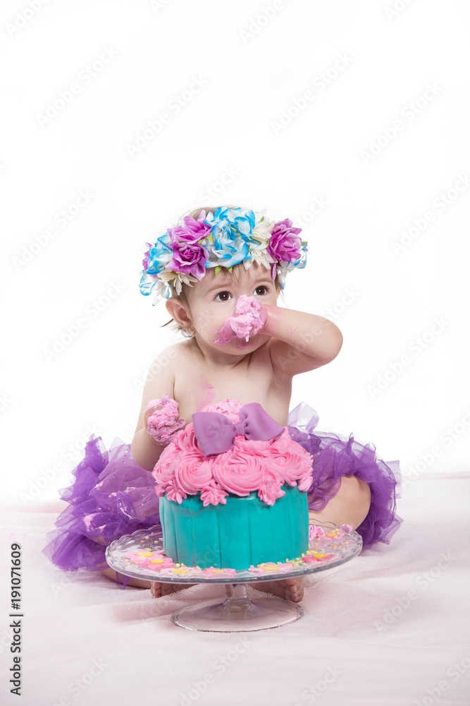 Cute happy baby girl with blue eyes is tasting the cake and purple butter icing on her sticky fingers from her first birthday cake smash while sitting on a white background . Cute happy baby girl with blue eyes is tasting the cake and purple butter icing on her sticky fingers from her first birthday cake smash while sitting on a white background .