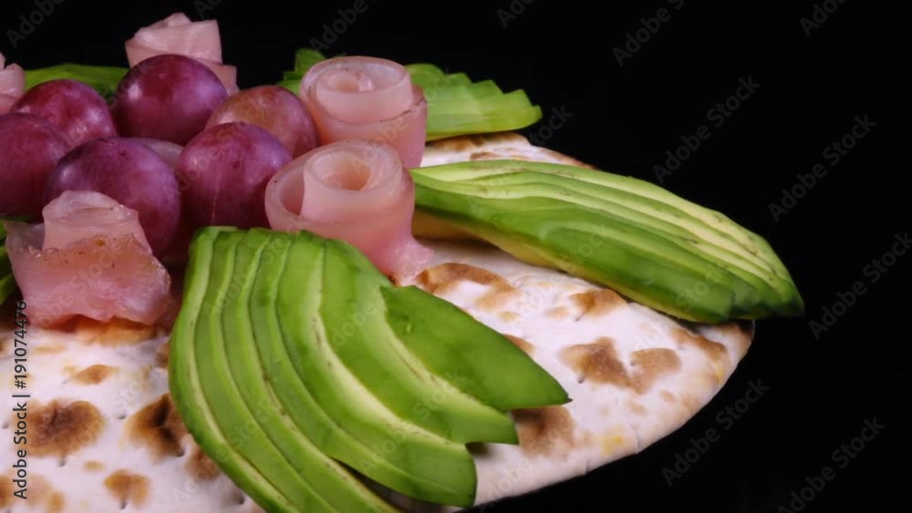 Homemade tasty pizza with avocado, grape and ham rotating against black background in 4K.
Homemade tasty pizza with avocado, grape and ham rotating against black background in 4K.
