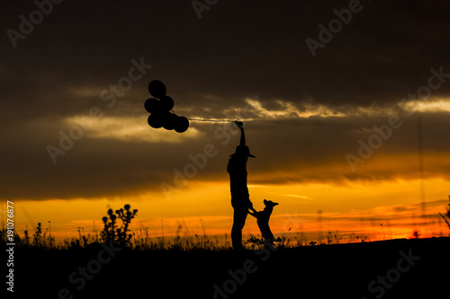 silhouette of a young woman and her dog at sunset. Outdoors. She is holdings balloons. Love for animals concept